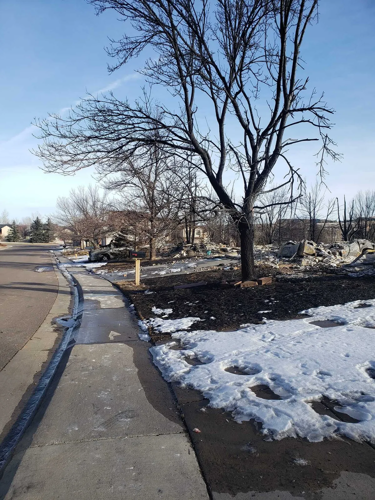 Picture of a sidewalk in Boulder County with a burned car and burned homes toward the right of the picture.