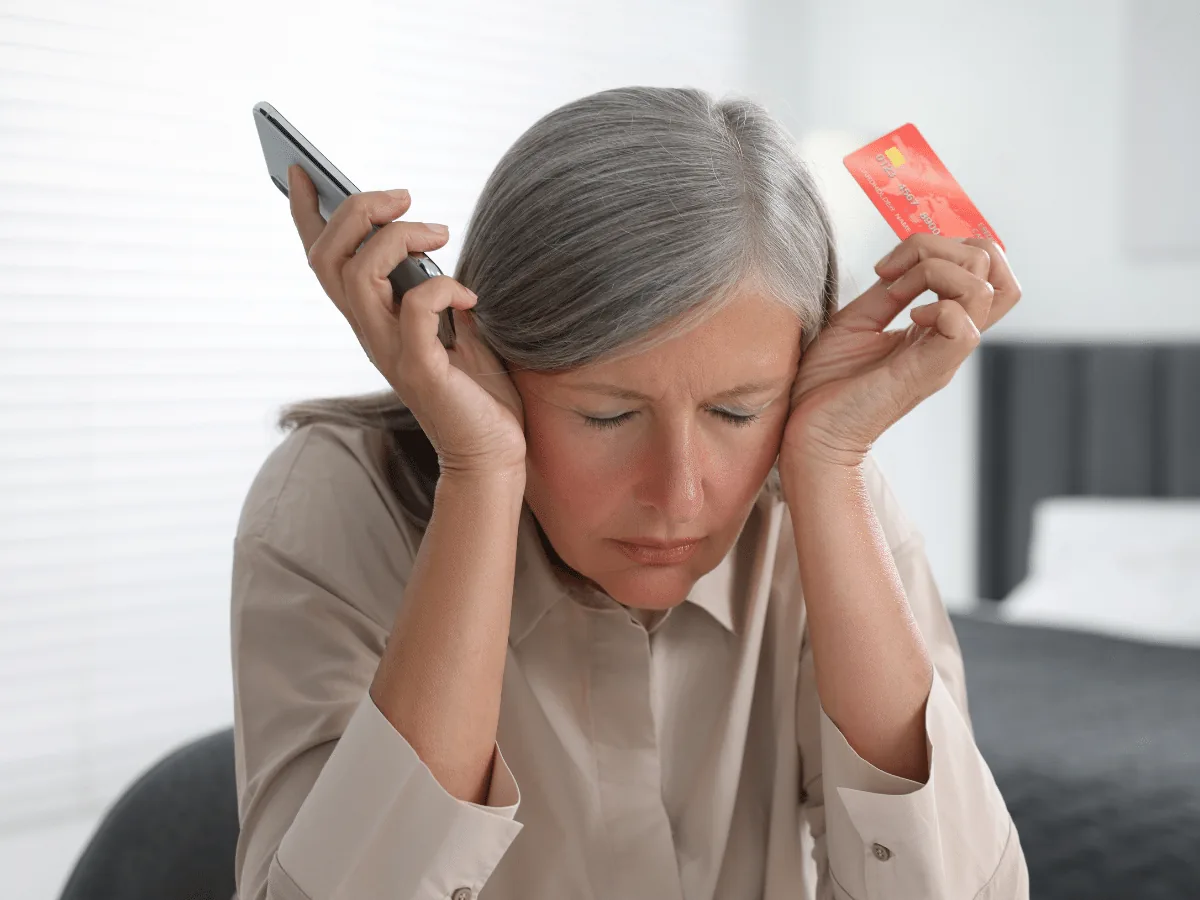 A lady with gray hair holds her phone and credit card looking frustrated by an online air duct cleaning scam.